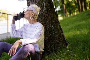 A woman drinking water outside under a tree after exercising.